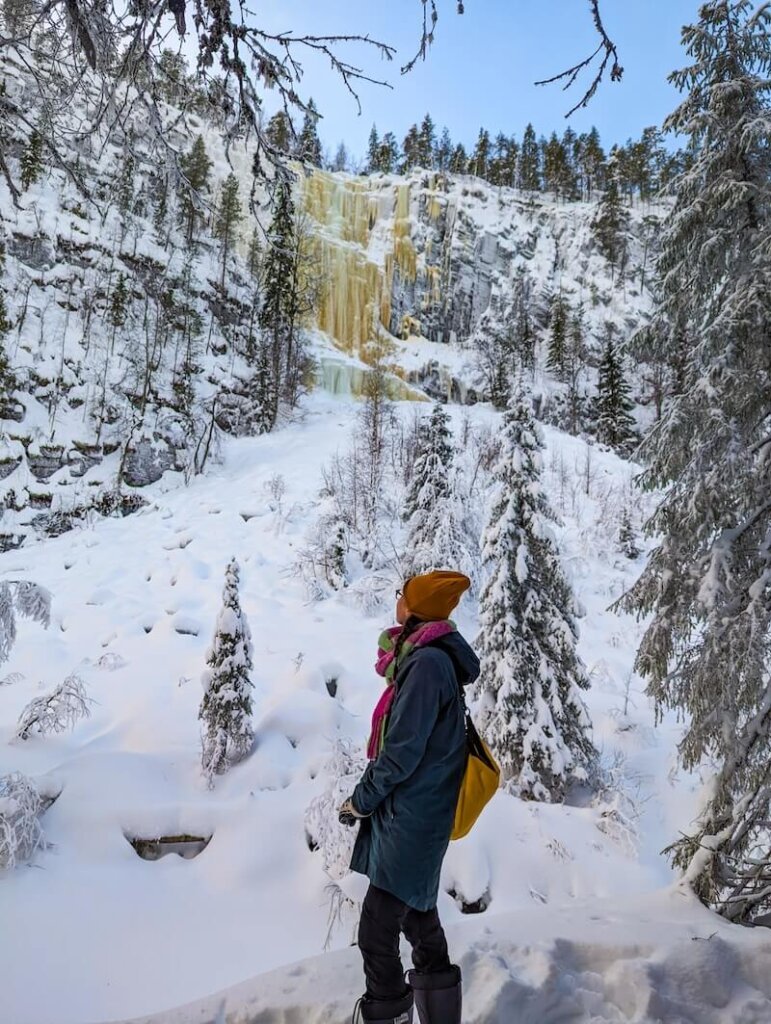 Allison Green wearing winter clothes (jacket, hat, scarf, boots) and yellow dry bag, standing in front of the icefall called Brown River in Korouoma Canyon in Finnish Lapland