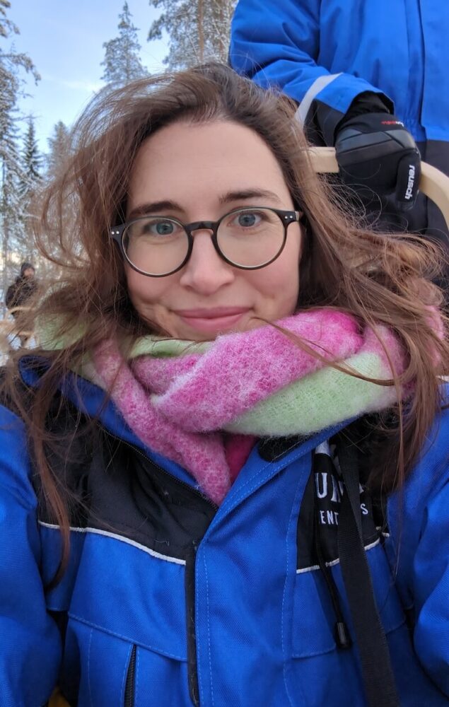 Allison smiling and sitting in the fog sled as the other traveler leads the dog sled and makes sure all are safe