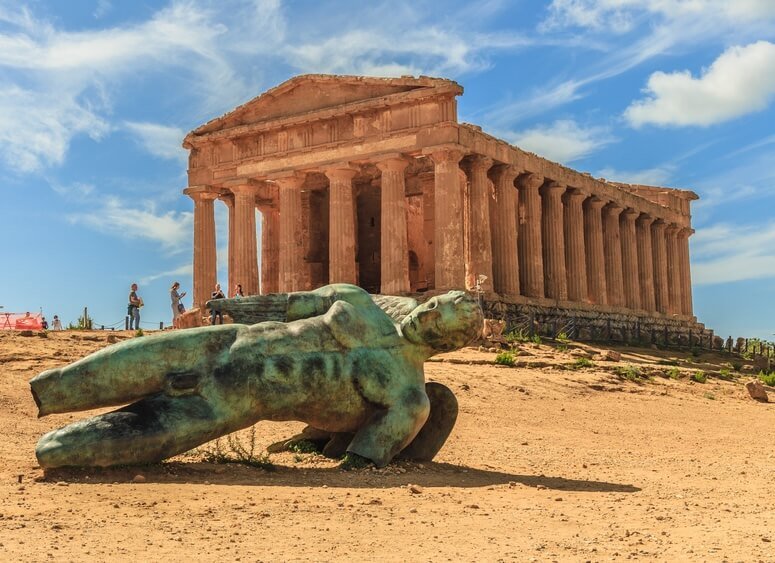 a bronze figure sculpture in front of one of the temple ruins of an ancient greek city in the sicilian town of agrigento