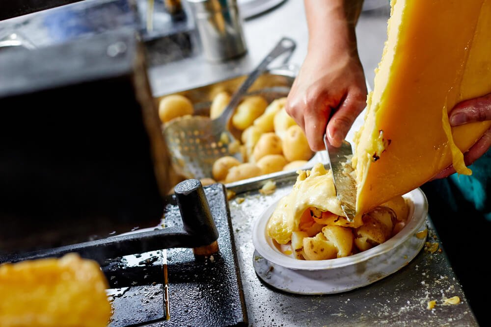 Someone cutting a melted slab of raclette over a dish of boiled potatoes and other deliciousness