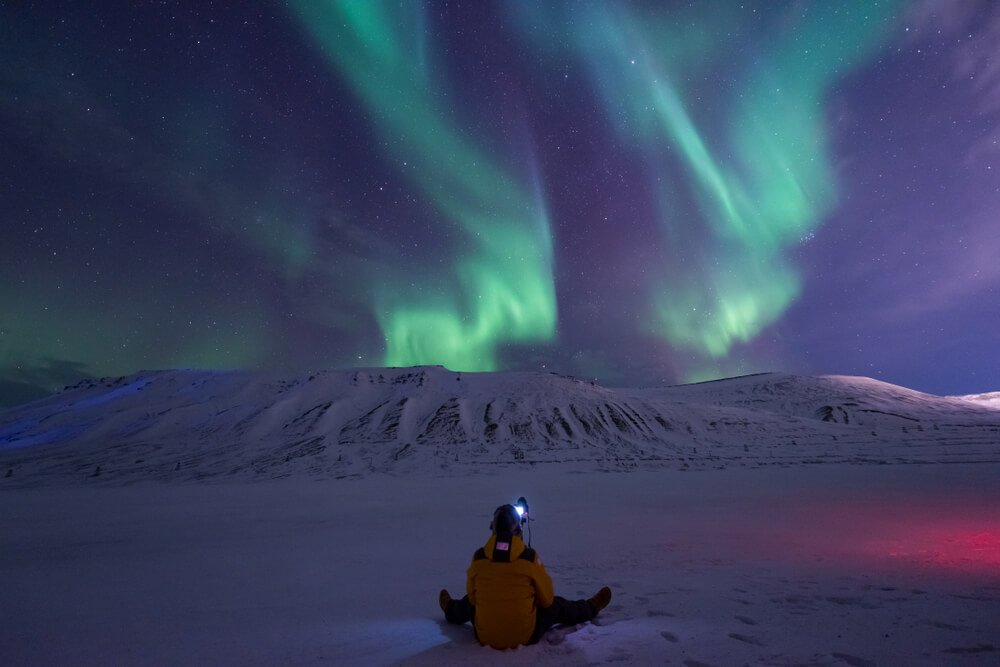Person with a headlamp on, sitting down in the snow, looking up at the Northern lights as they shoot around overhead
