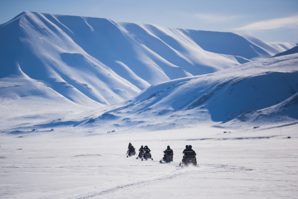 A group of five people snowmobiling through a valley with huge mountains surrounding them as they traverse the rugged Svalbard landscape