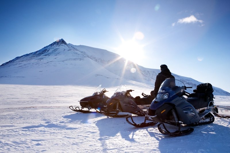 Three snowmobiles and a winter landscape with mountain, and the sun is starting to set behind the mountain despite it being quite bright outside, there is one person in the shot too.