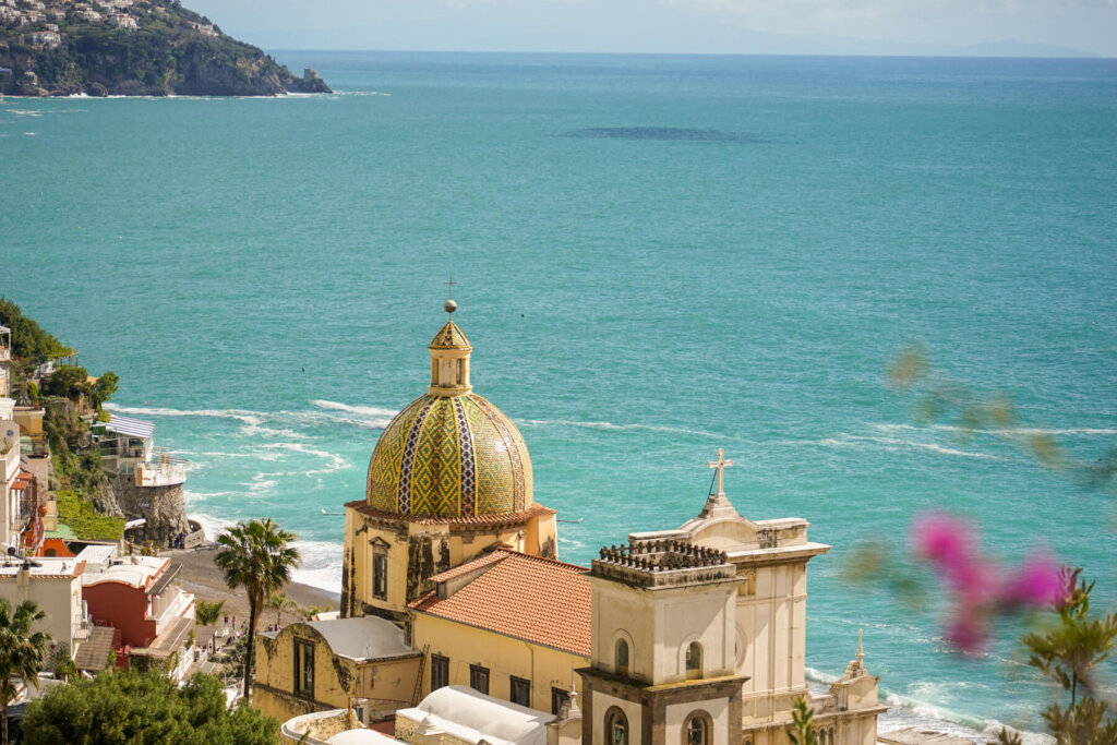 View of the sea of Positano in march with the famous tile-roof church and pink flowers in the foreground and a beautiful landscape of the sea and coast