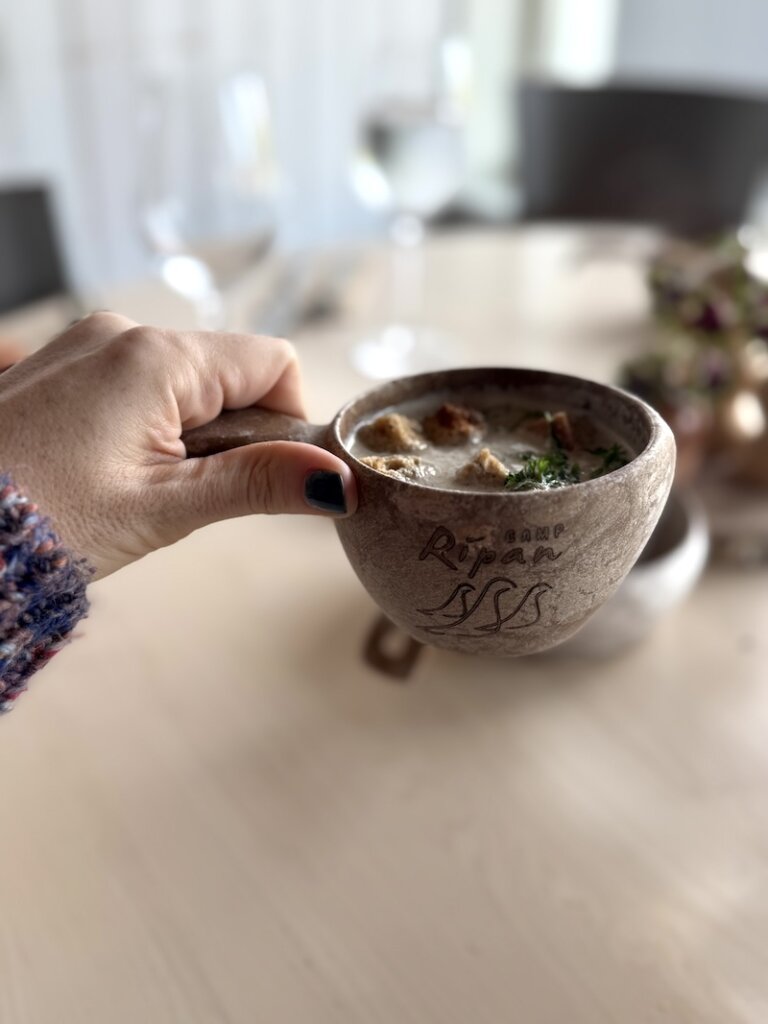 Allison's hand at the restaurant at Camp Ripan, called ripan kitchen, holding a cup of mushroom soup in a traditional wooden cup