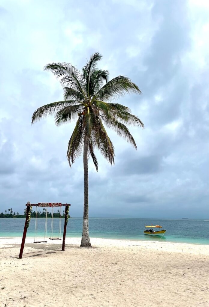 Swingset that says Guna Yala, sandy beach, palm tree, cloudy sky and boat docked in a shallow area