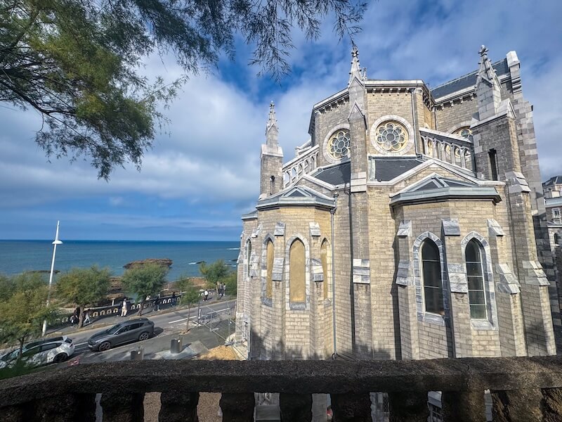 Gray stone church with two rose windows and three spires and lots of masonry detail, with tree, and view of the Atlantic ocean and a promenade behind it.