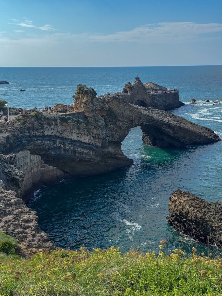 Foreground of yellow wildflowers overlooking a rugged coastal cliff with bright blue water below. A manmade bridge connects the mainland to a rocky outcrop, where several people are walking and enjoying the view.