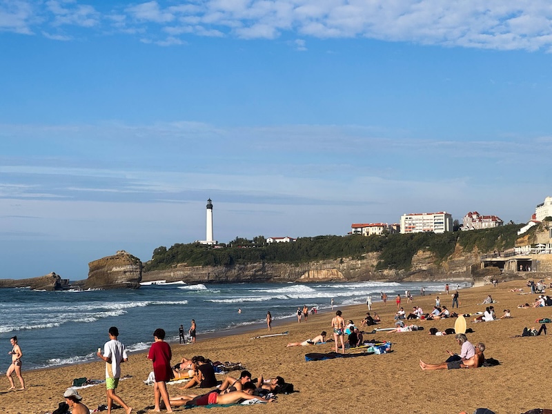 The main "grand plage" of Biarritz, the main beach, with lots of crashing waves, people enjoying the sand, towels laid out, with a view of a lighthouse in the cliff in the distance
