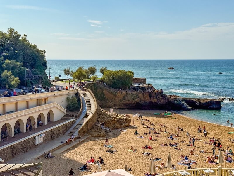 Sunny day at a horseshoe-shaped beach in Biarritz, with colorful umbrellas and towels scattered across the sand. Remnants of its old port remain visible, including stone archways and sloped stairways leading down to the water.