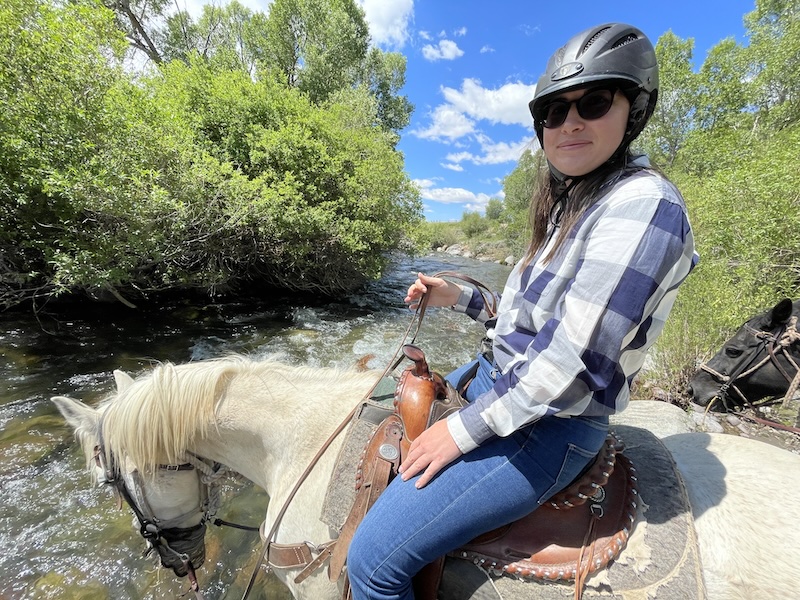 Allison on a horse in "blue sky country" in wyoming with her horse drinking from the water of a river