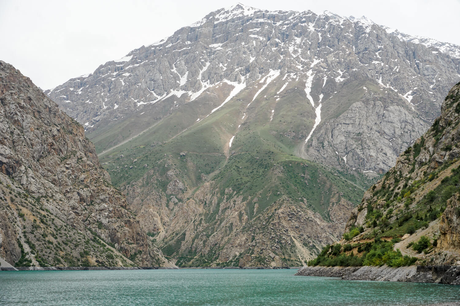 A mountain with snow covering it in little rivulets and a turquoise colored lake with clouds overhead