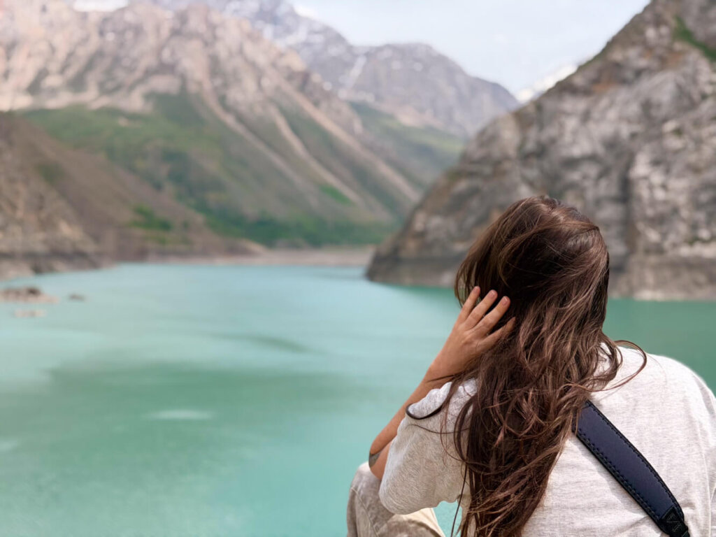 Allison holding her hair, blowing in the wind as the mountains are at high elevation, while looking out at the 6th lake of Tajikistan's 7 Lakes